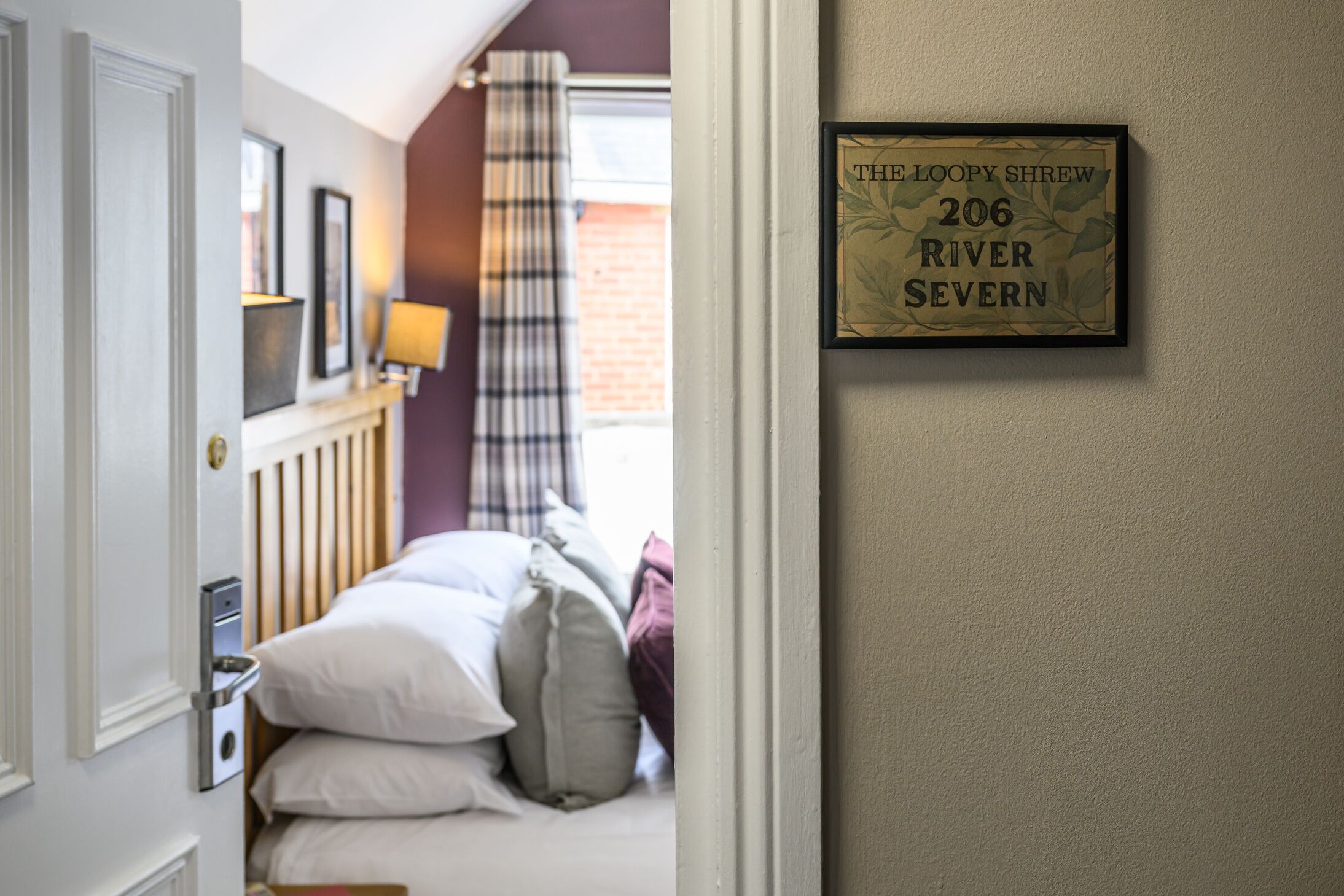 Une chambre à coucher avec un lit, des oreillers, une table de chevet avec une lampe, un cadre décoratif accroché au mur et une fenêtre avec un rideau.
