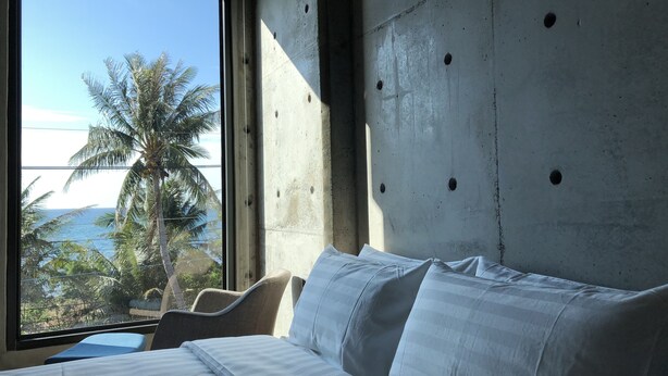 Ein Zimmer mit großem Fenster, einem Bett mit weißer Bettwäsche und einer Betonwand mit Löchern.