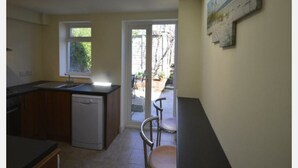 A compact kitchen with a dishwasher, a window, and a door leading to a small outdoor area with chairs. - Victorian Rose Cottage (Deal)