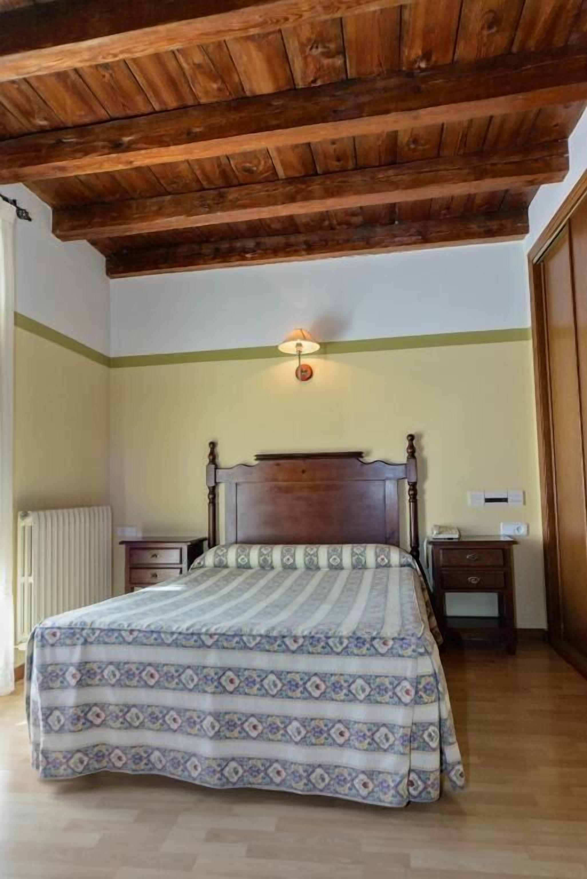 Ein Schlafzimmer mit Holzbalkendecke, einem Bett mit gemustertem Überwurf, einem Nachttisch und einer Wandlampe.