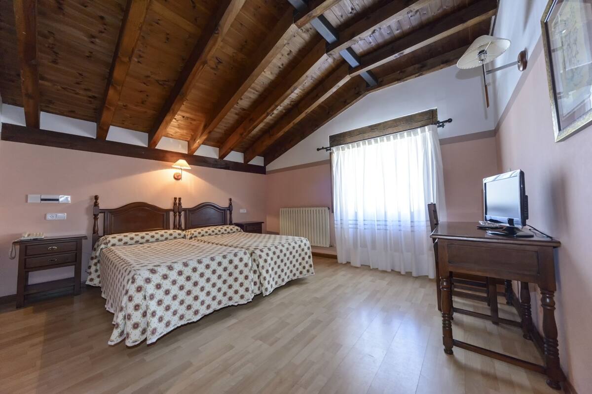 Un dormitorio con techo de madera, una cama con colcha estampada, un escritorio con computadora y una ventana con cortinas.