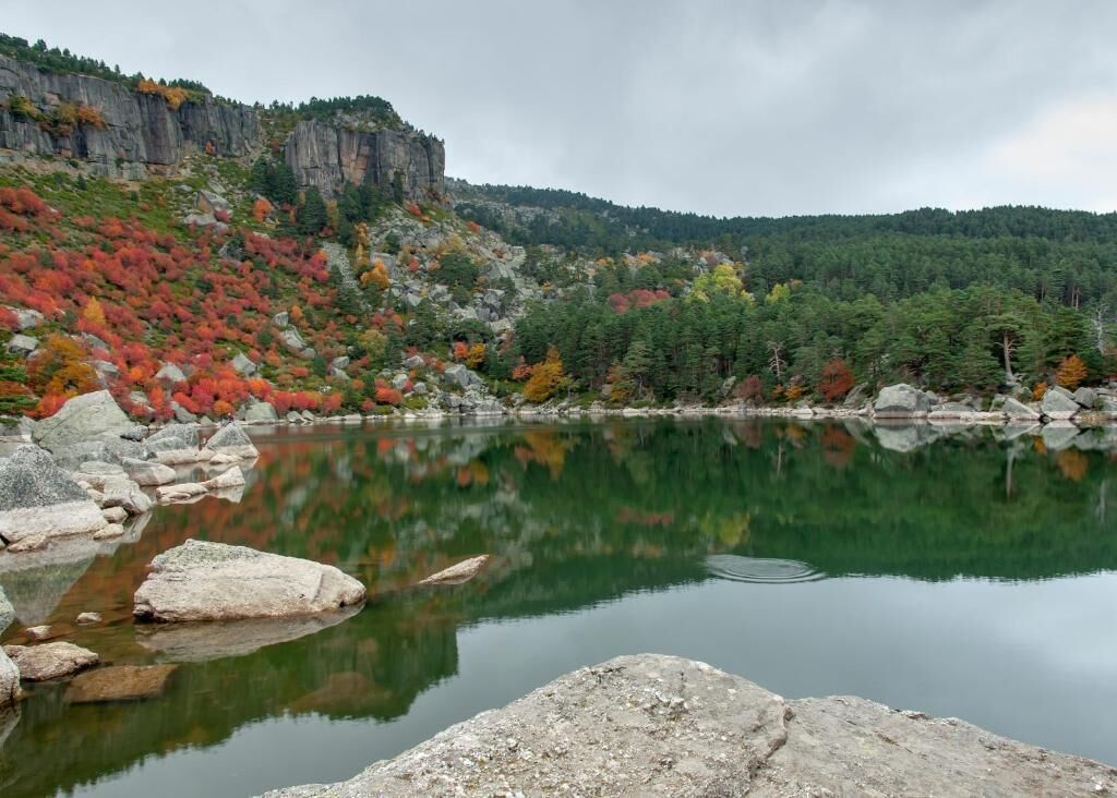 A calm lake with rocky shores and autumn-colored trees reflecting on the water.