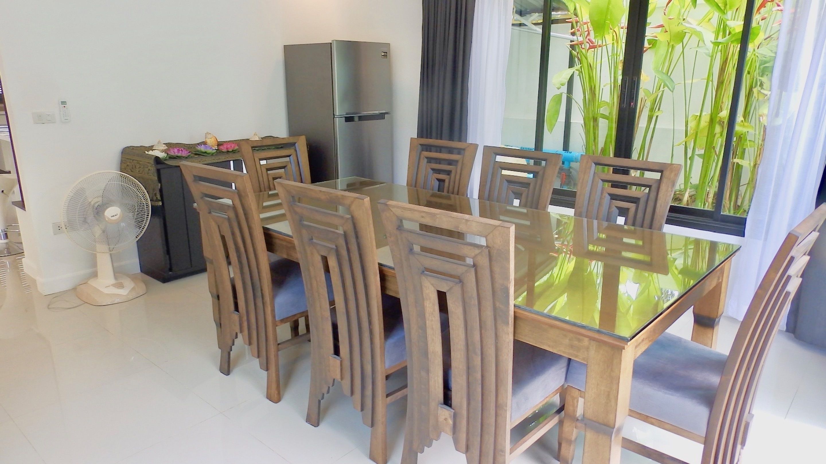 A dining area with a glass table and wooden chairs, a refrigerator, a standing fan, and a view of greenery through a window.