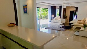 A modern living room with a sofa, a coffee table, and a TV. There is a glass door leading to a balcony with plants. - 3 Bedroom Private Pool Villa Flora (Koh Samui)