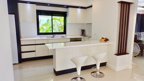 A modern kitchen with a white countertop, two bar stools, a window with a view of greenery, and a fruit bowl on the counter. - 3 Bedroom Private Pool Villa Flora (Koh Samui)