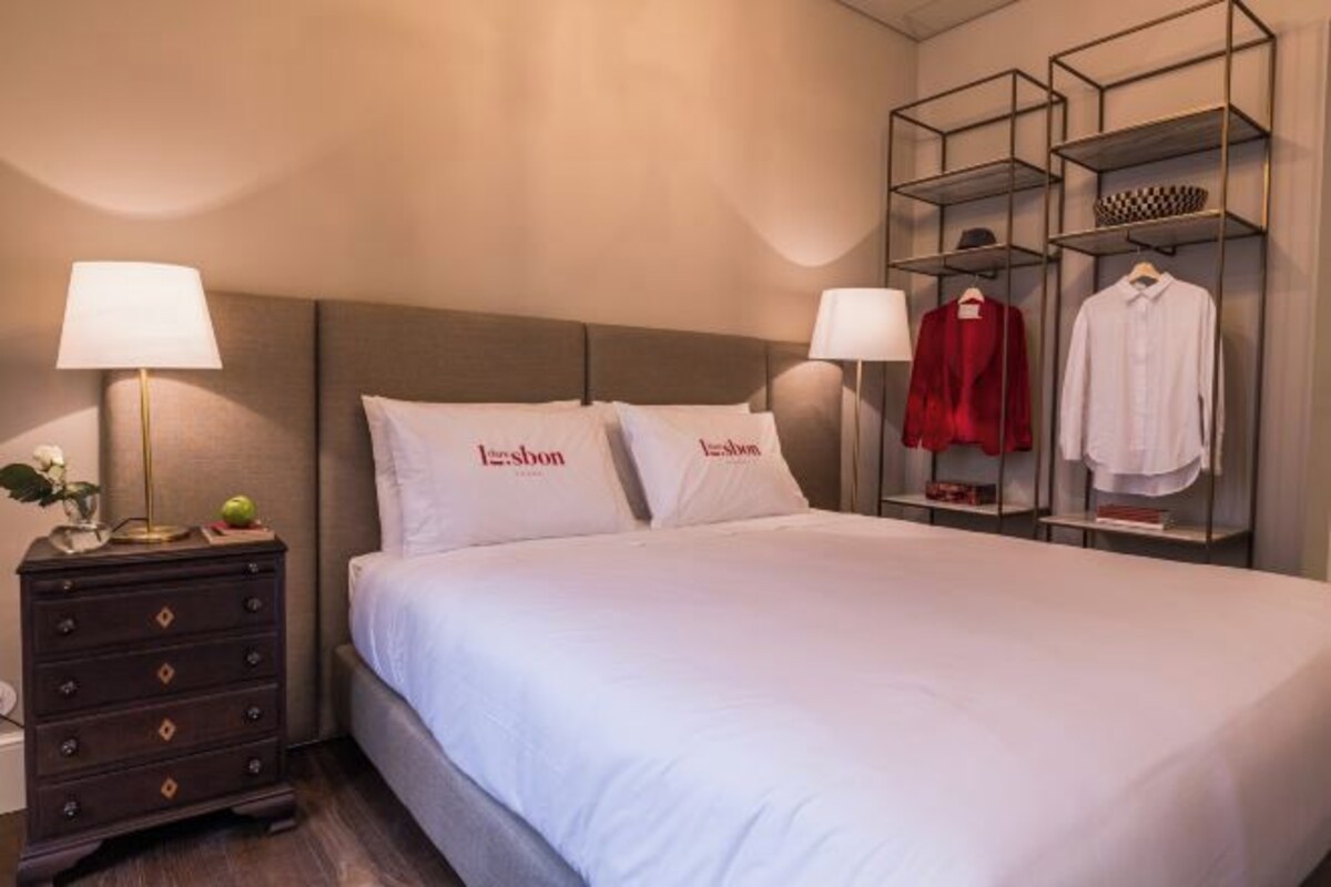 Una habitación de hotel con cama, dos lámparas de noche, cómoda y perchero.