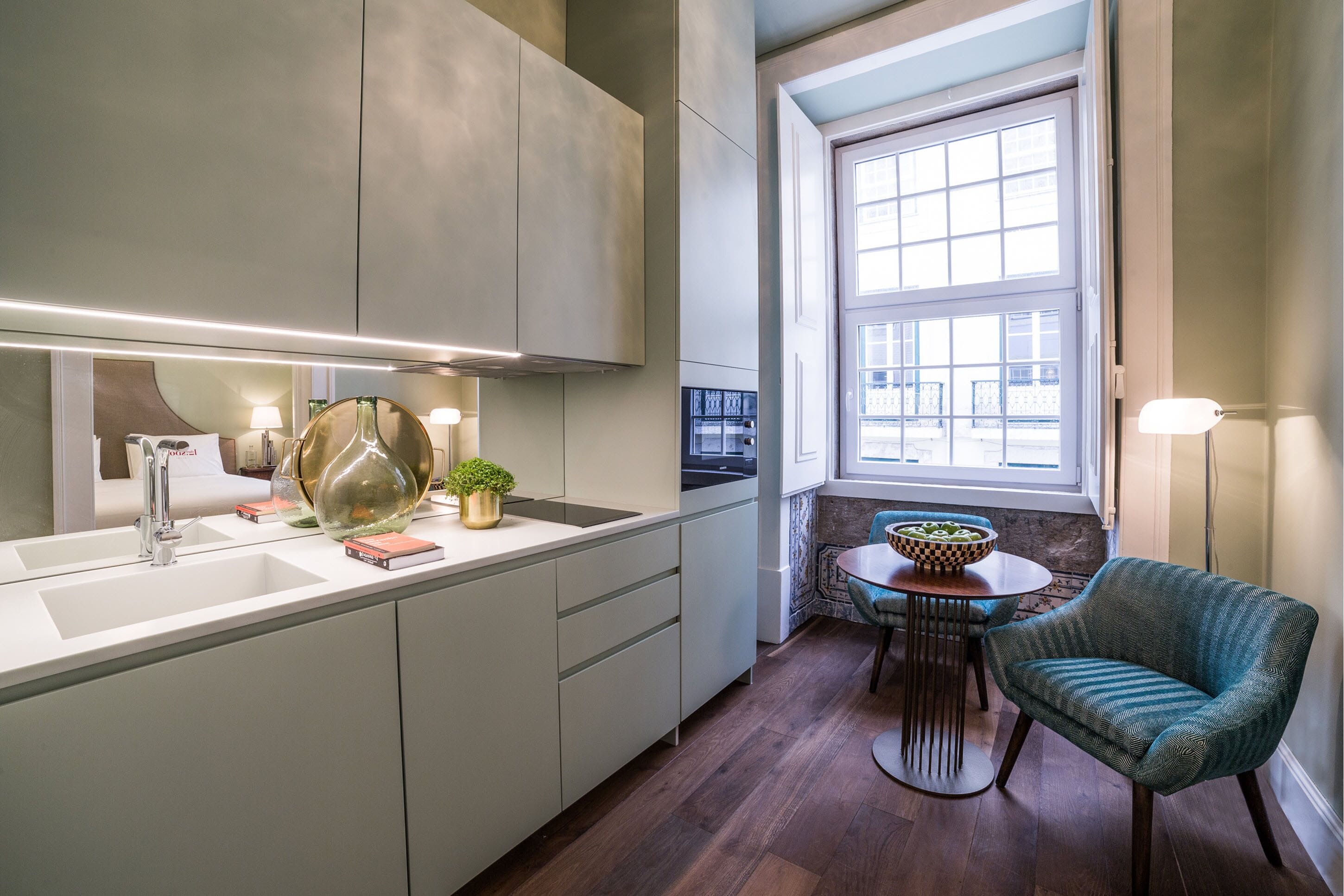 A modern kitchen with a large window, a dining area with a round table, and a teal armchair.