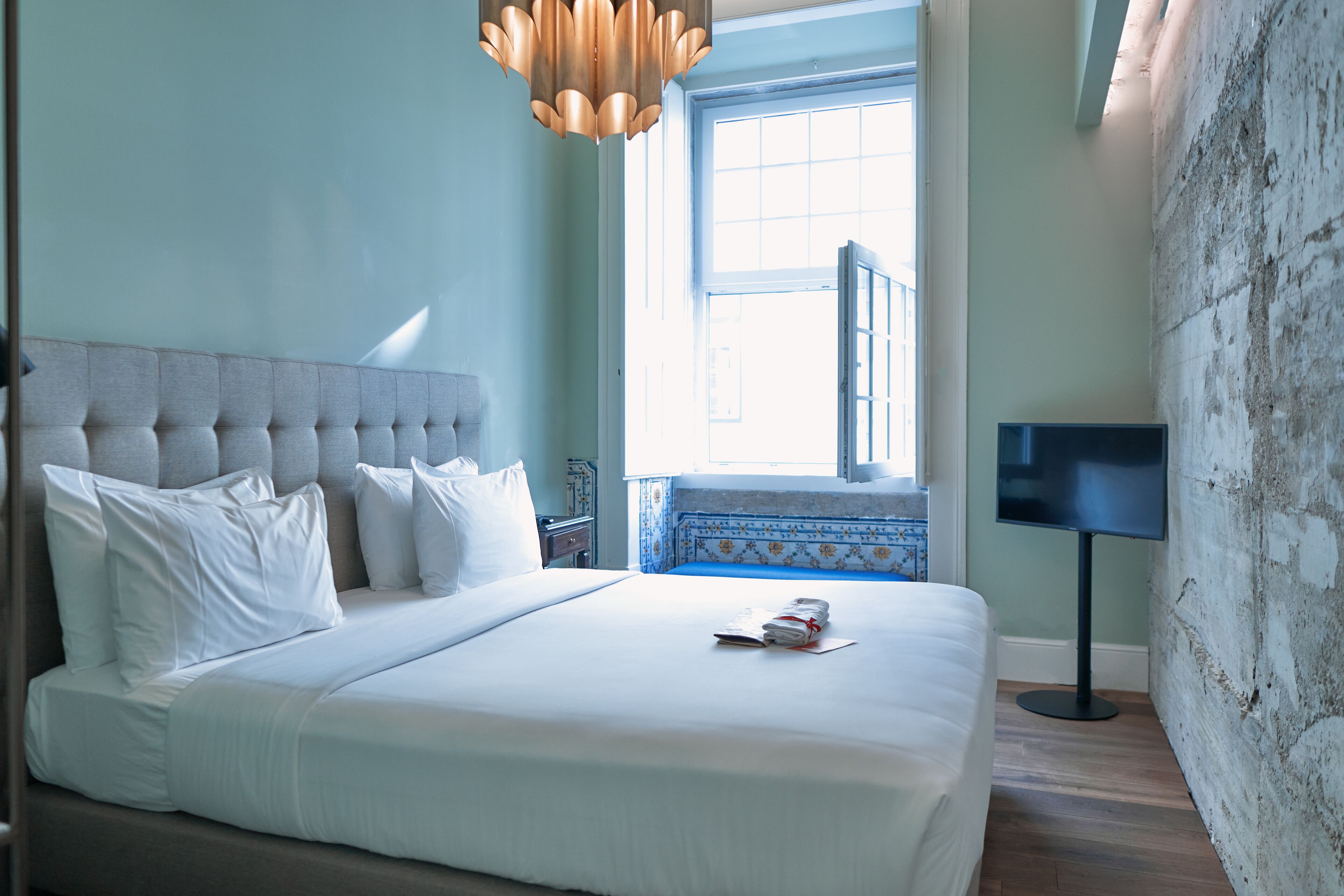 A neatly made bed with white linens and pillows, a modern chandelier, a television on a stand, and a large window with a view of the outside.