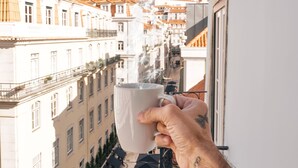 Une main tenant une tasse blanche, avec en arrière-plan une rue bordée d’immeubles et de terrasses.