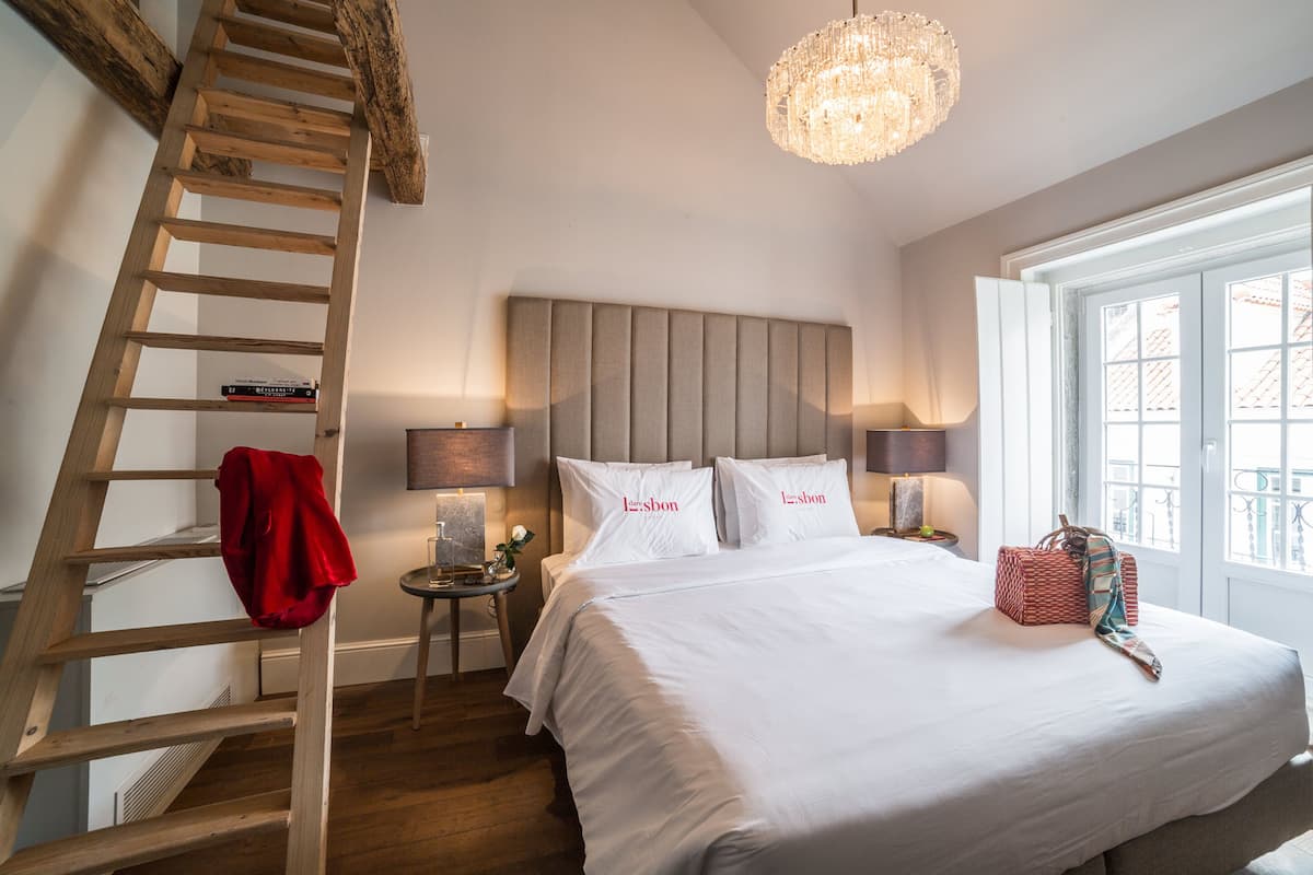 Une chambre avec une échelle en bois, un lit avec une literie blanche, un lustre et une fenêtre donnant sur un beau paysage.