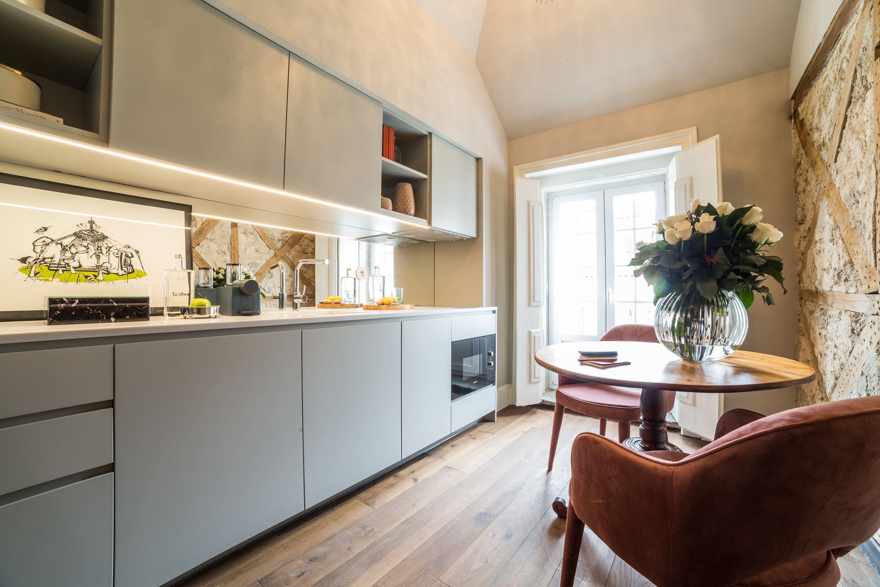A modern kitchen with grey cabinets, a marble countertop, and a dining area with a round table and red chairs.