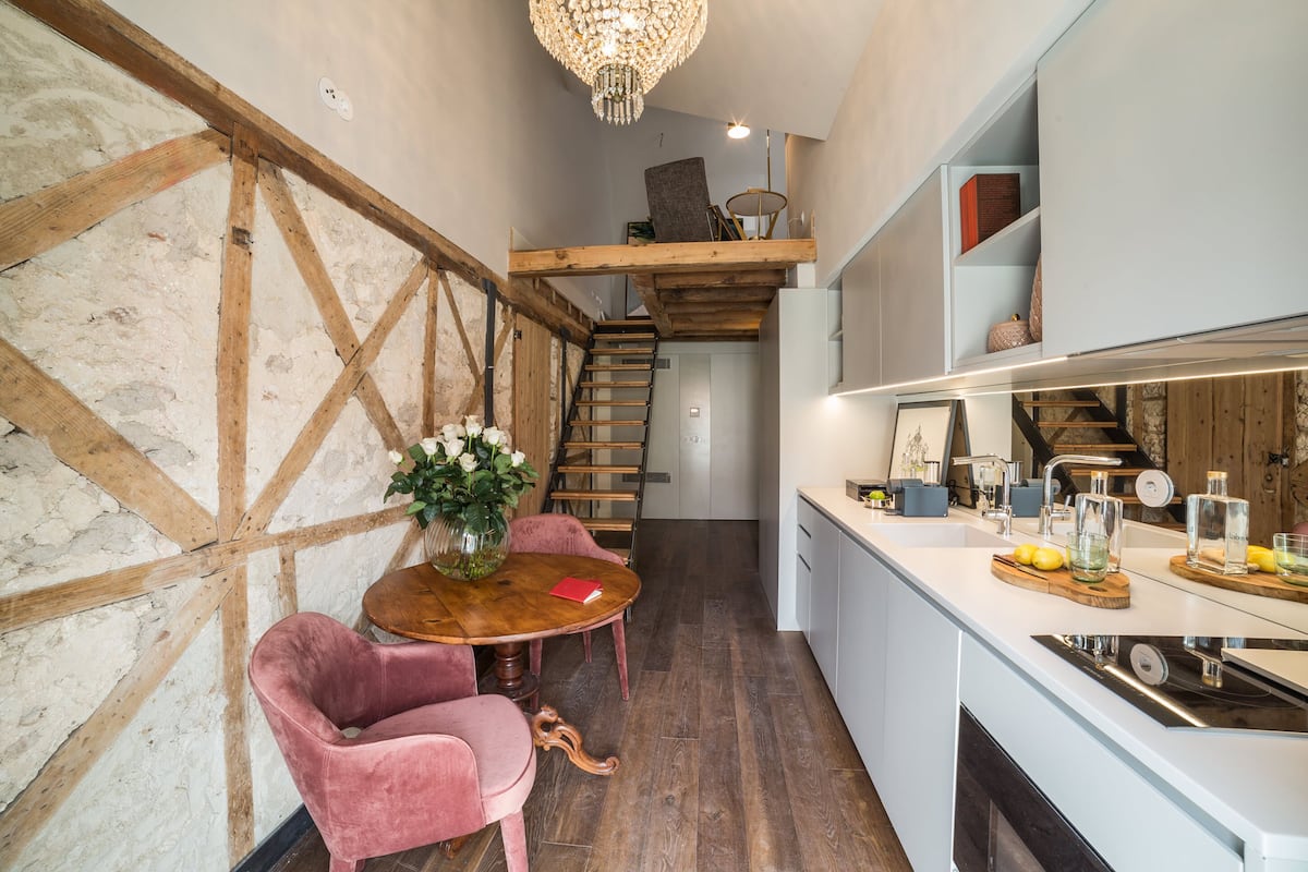 Une cuisine moderne avec un escalier en bois, un coin repas agrémenté d’une chaise rose et un lustre.