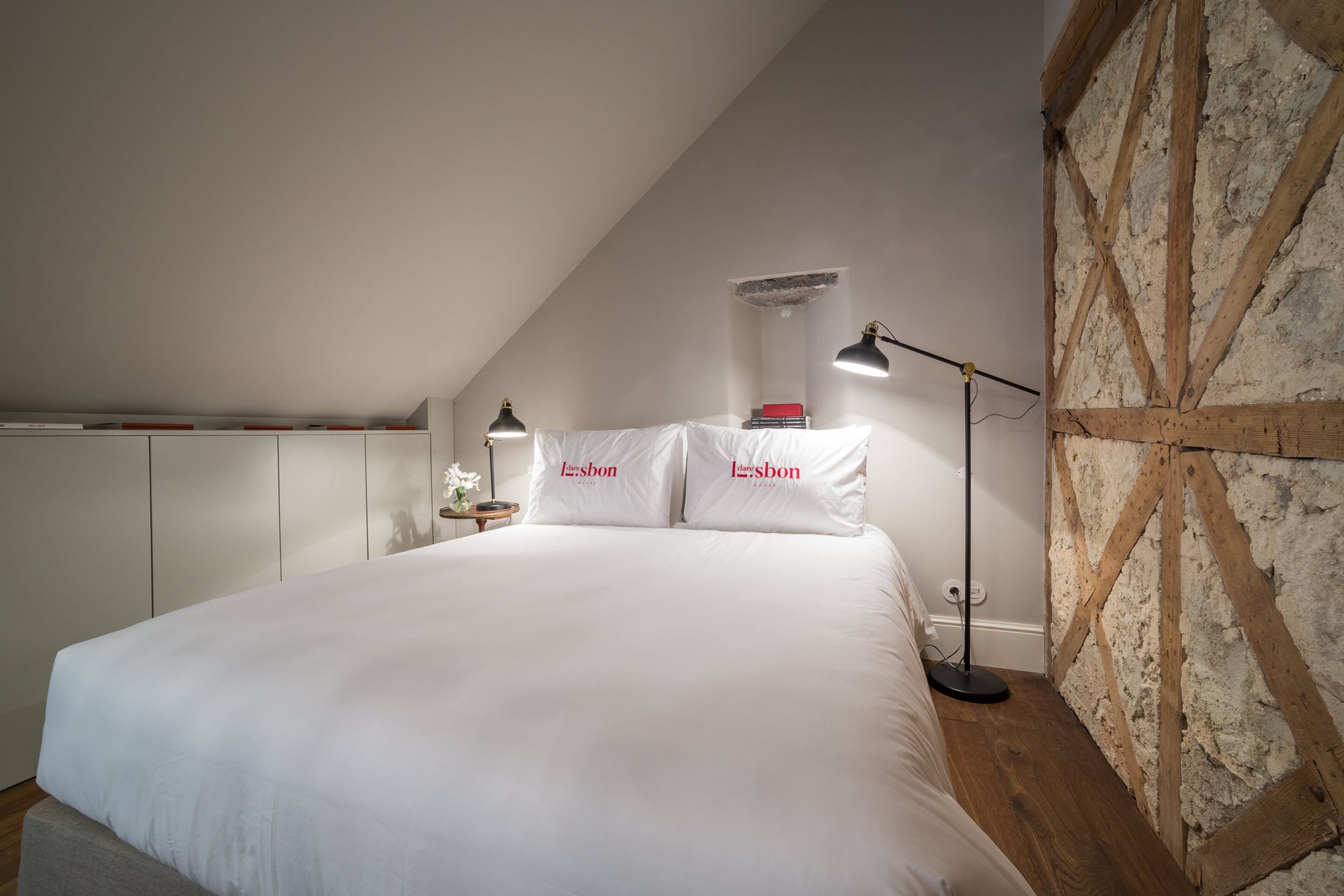 A bedroom with a large bed, white bedding, two bedside lamps, a nightstand, and a stone wall.