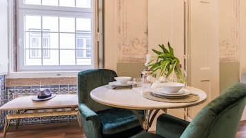 Un comedor con una mesa redonda dispuesta para dos personas, dos sillas de terciopelo verde, un sombrero blanco sobre un banco y una planta en maceta sobre la mesa.