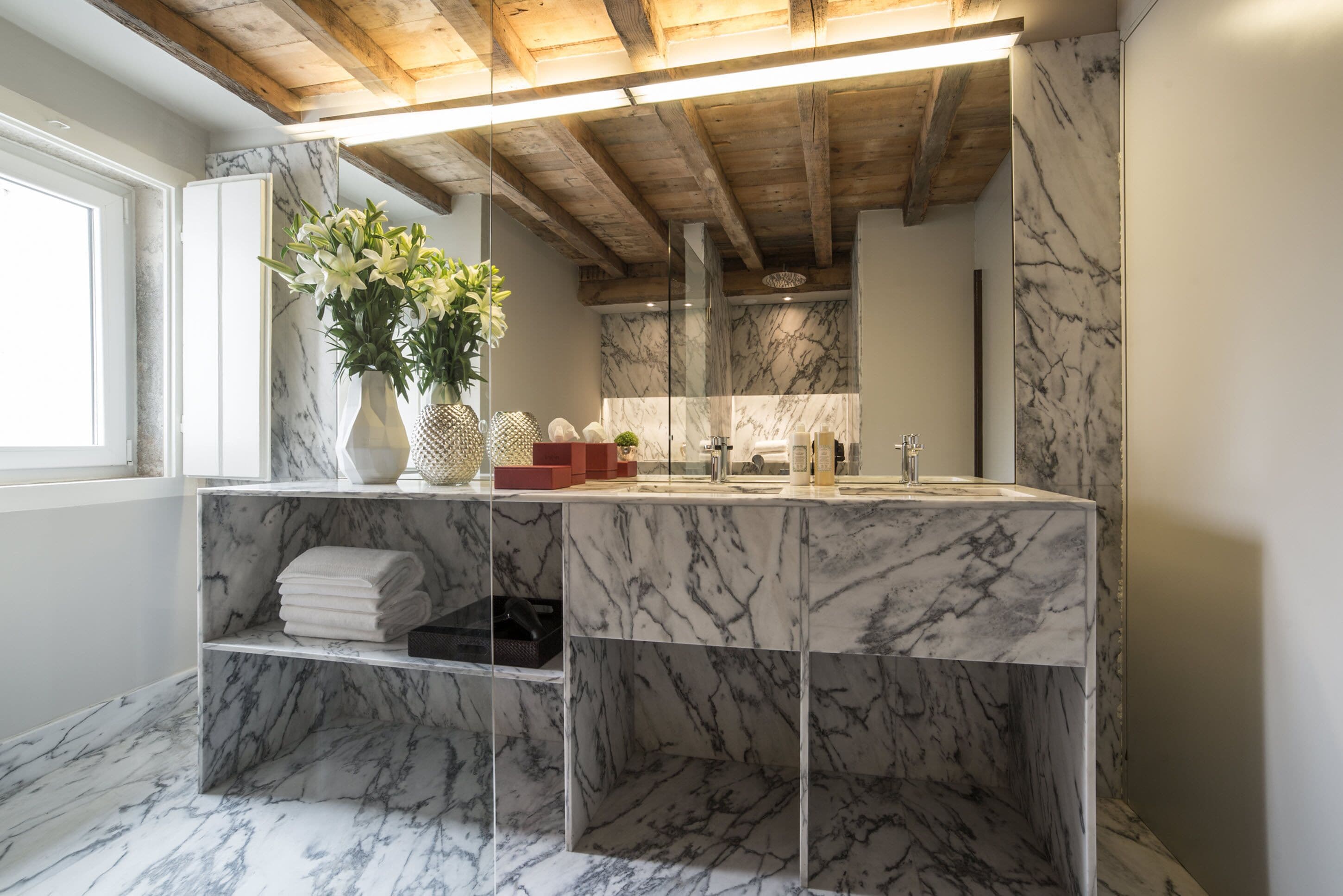 Un bagno con pavimento in marmo, scaffali, soffitto in legno e un grande specchio.