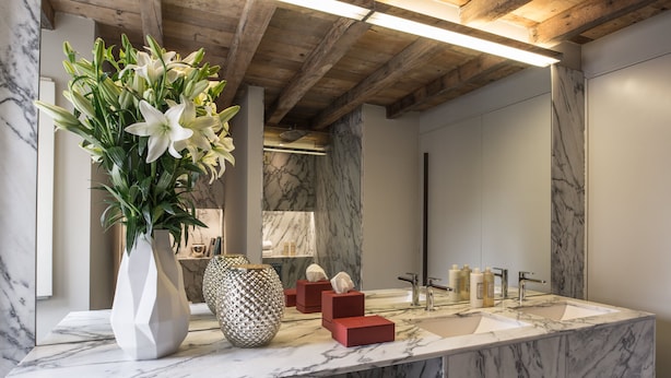 Une salle de bain avec des comptoirs en marbre, un grand miroir, un vase rempli de fleurs blanches et un plafond en bois.