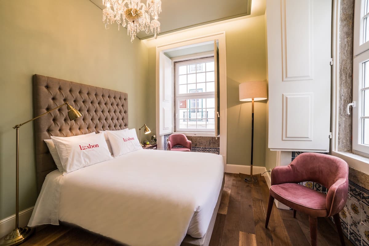 Un lit bien fait, doté d’une tête de lit capitonnée, une chaise rose et un lustre.