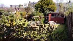 A fenced backyard with a wooden swing set, a red shed, and a large tree. - Fishermen's Cottage (Deal)