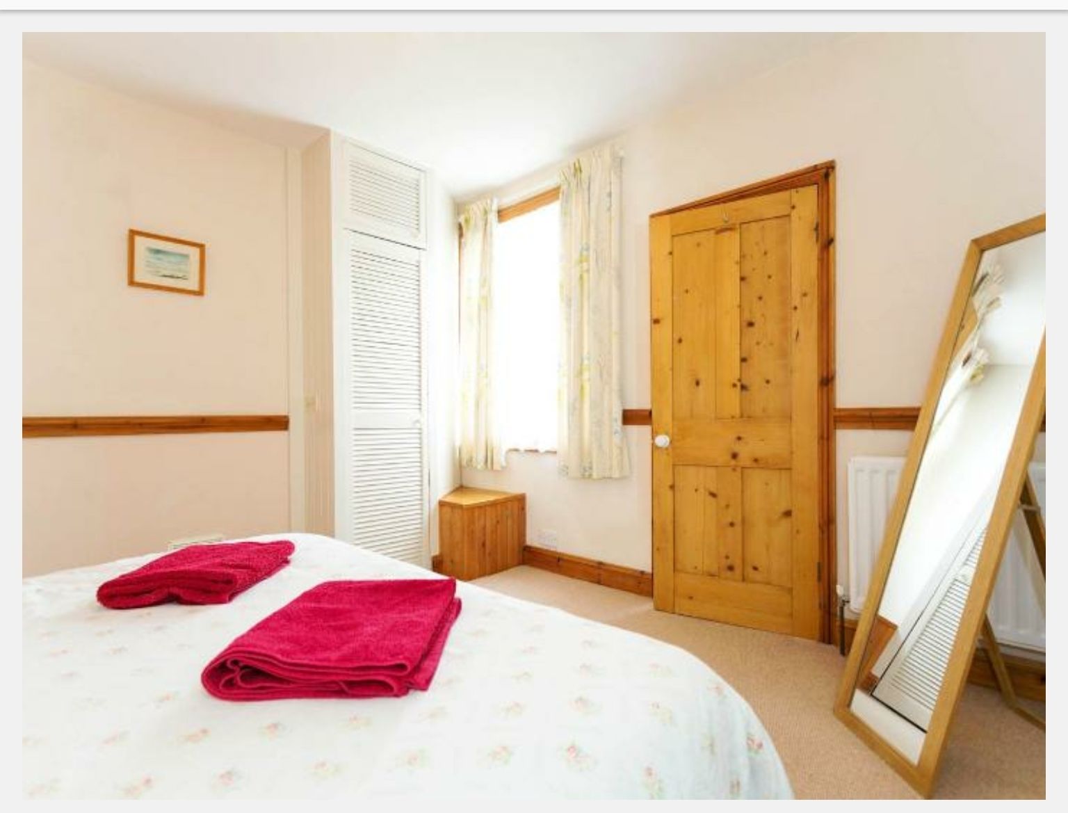 Un dormitorio con una cama, dos toallas enrolladas, una puerta de madera, una ventana con cortinas, un taburete de madera pequeño y un espejo en marco apoyado contra la pared.