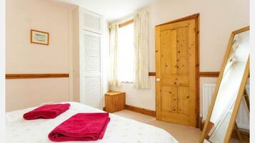 Un dormitorio con una cama, dos toallas enrolladas, una puerta de madera, una ventana con cortinas, un taburete de madera pequeño y un espejo en marco apoyado contra la pared.