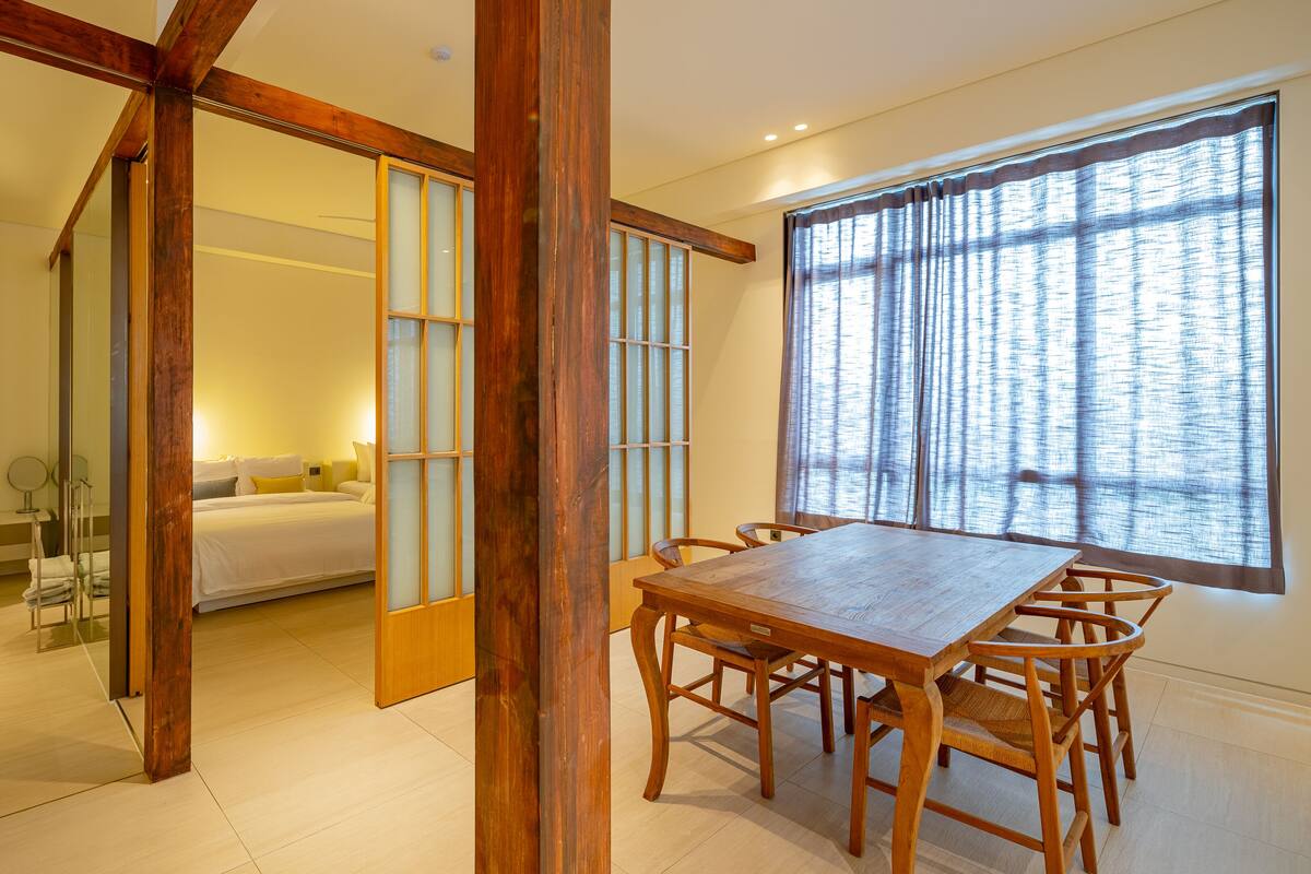 A hotel room with a wooden dining table, chairs, a large window with curtains, a bed with white bedding, and a bathroom visible through an open door.