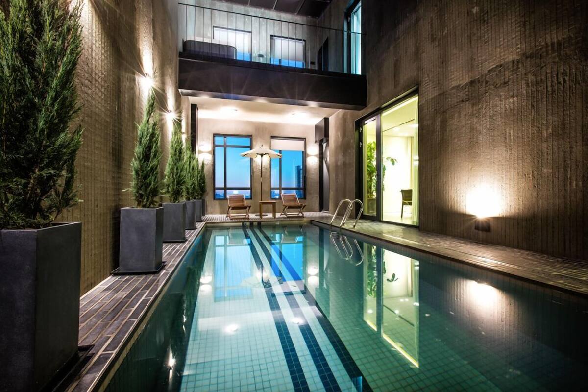 An indoor swimming pool with a modern design, featuring a clear pool, wooden deck, and seating area with chairs.