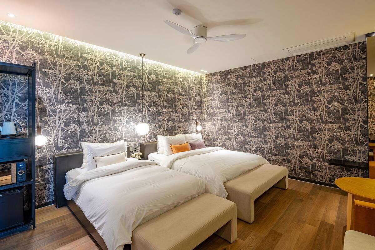 A modern hotel room with two beds, a ceiling fan, a small table, and a mirror.