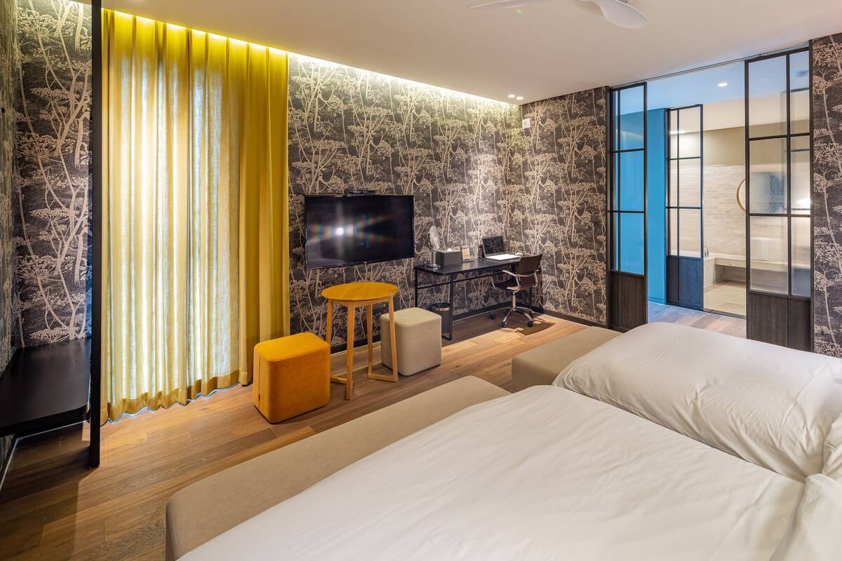 A modern hotel room with a large bed, a desk with a computer, a TV, and a bathroom visible through a glass door.