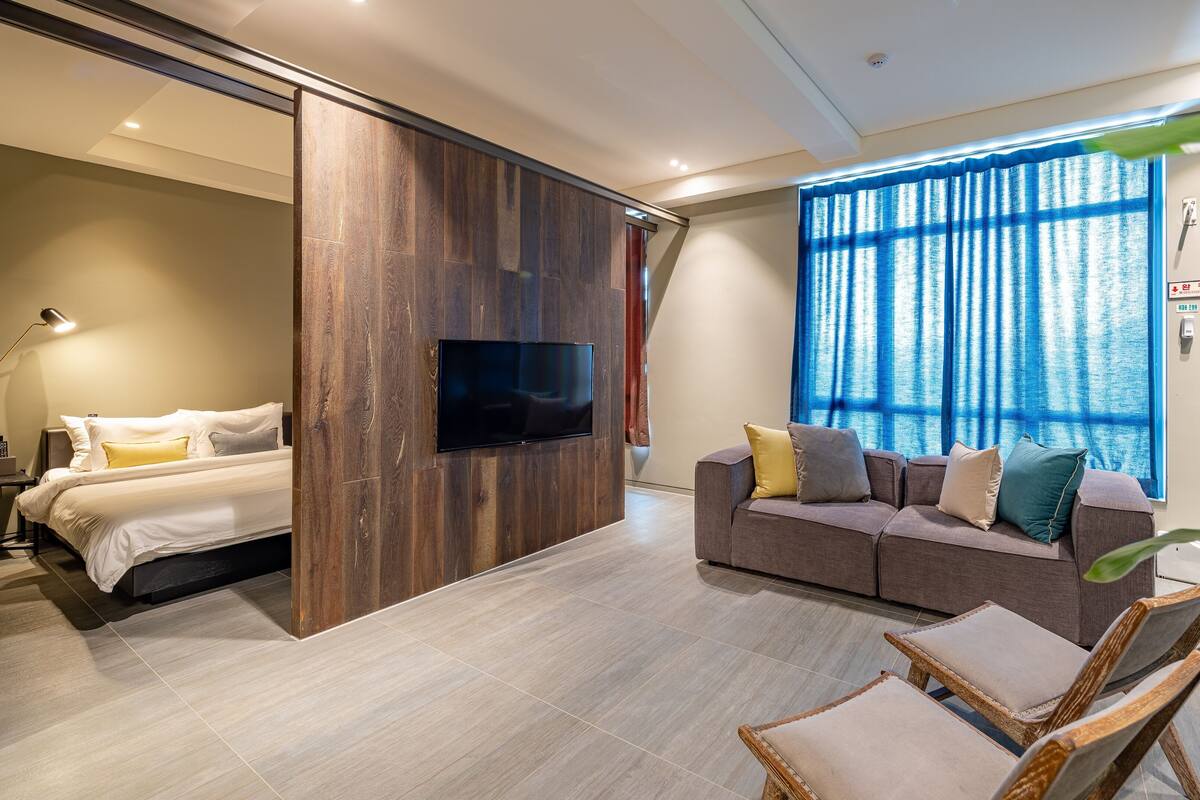 A modern hotel room with a bed, a sofa, a TV, and a large window with curtains.