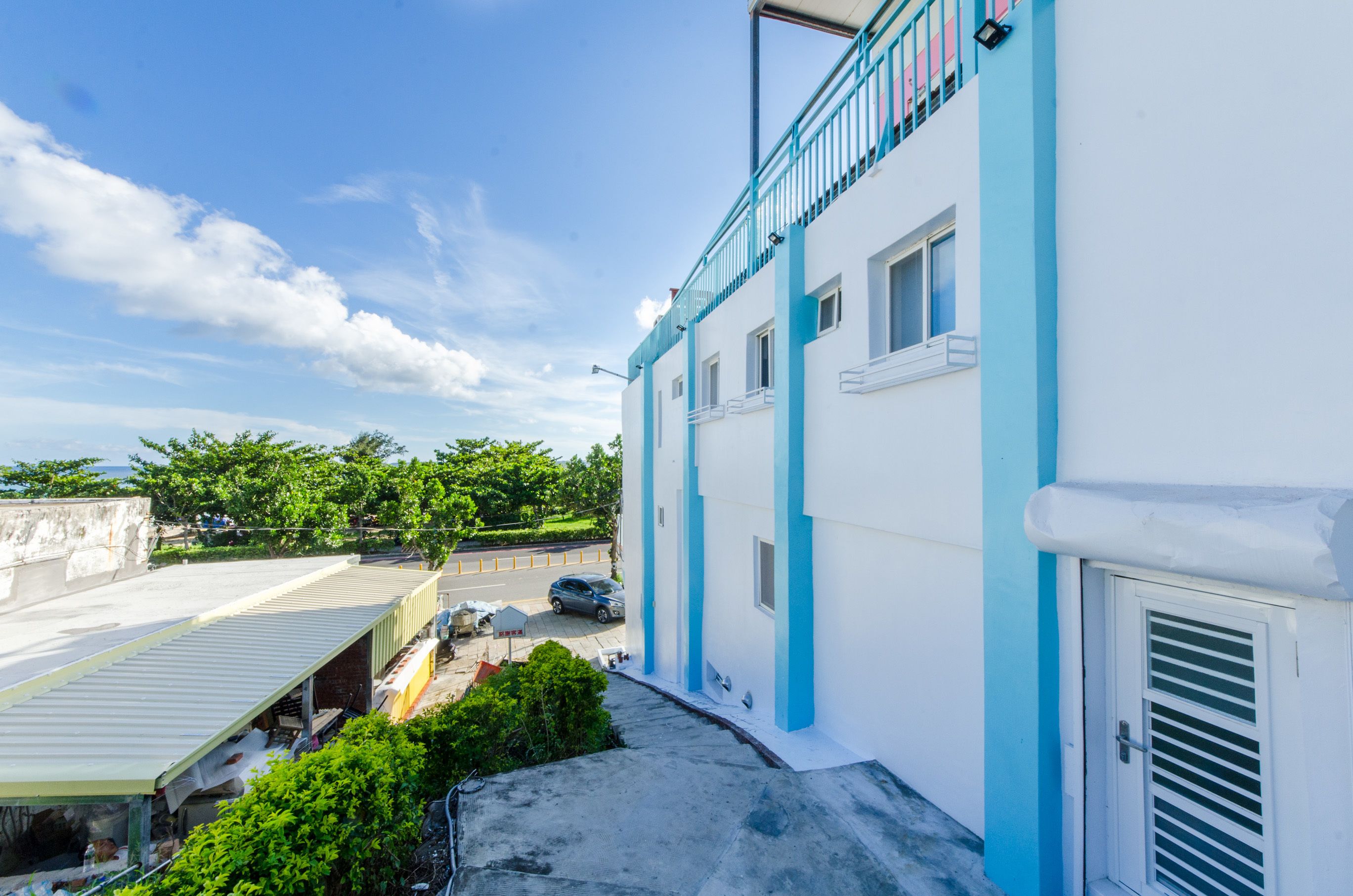 Um edifício com fachada azul e branca, uma varanda e vista para uma rua com carros estacionados e vegetação.