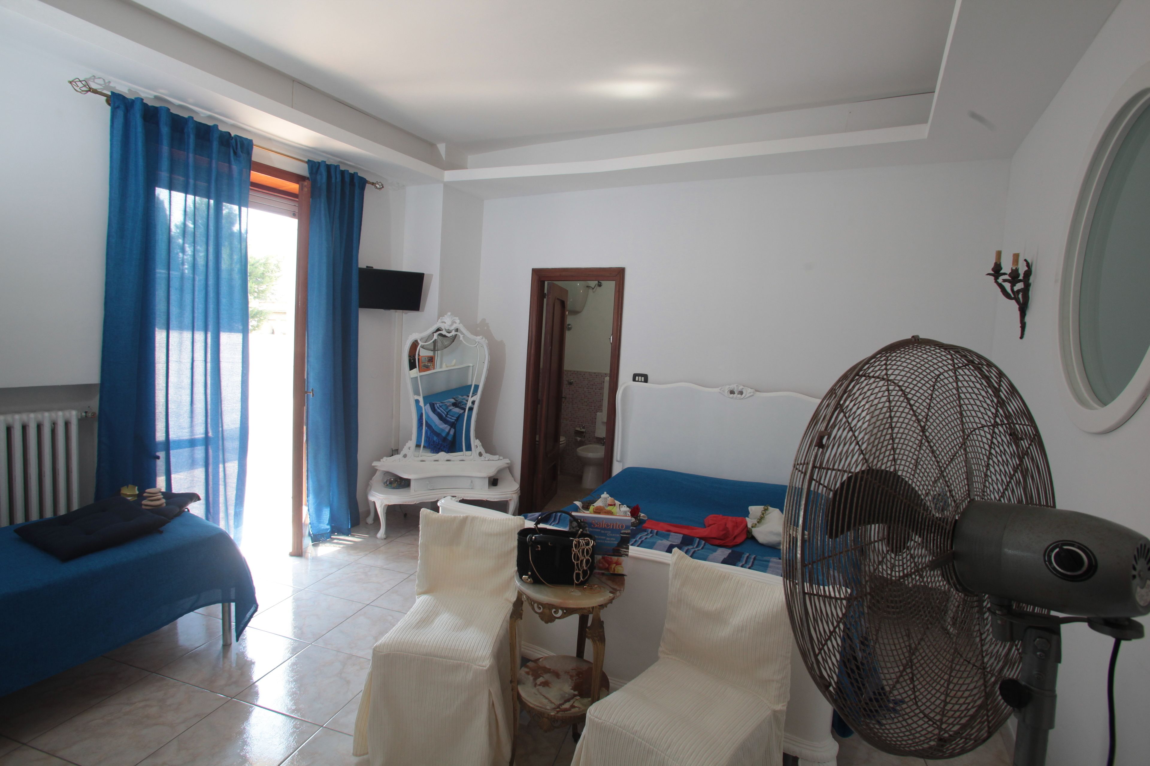 Una habitación con una cama, una silla, una mesa, un ventilador y una ventana con cortinas.