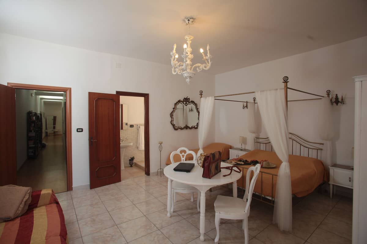 A bedroom with a bed, a table with chairs, a chandelier, and a bathroom visible through an open door.