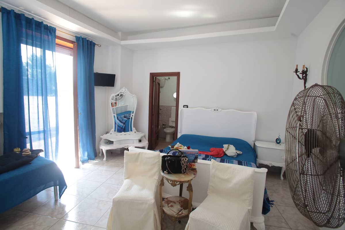 A bedroom with a bed, a chair, a small table, a fan, and a window with curtains.