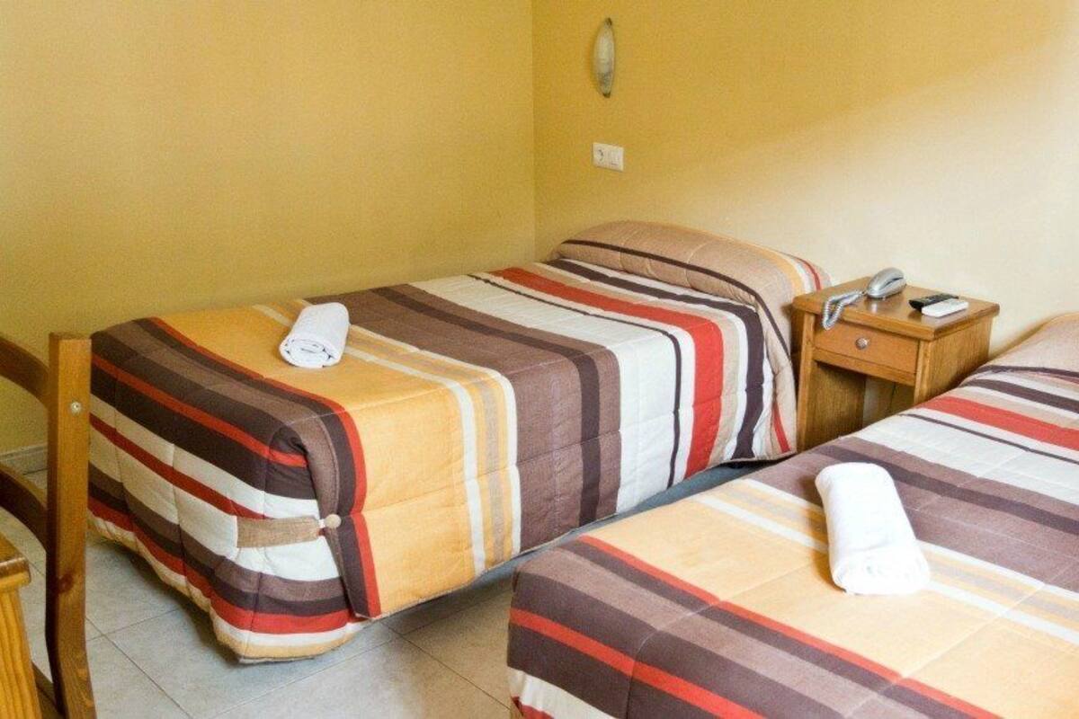 Una habitación de hotel pequeña con dos camas individuales, cada una con ropa de cama a rayas y una toalla enrollada. Hay una mesita de noche de madera con un teléfono y un enchufe de pared.
