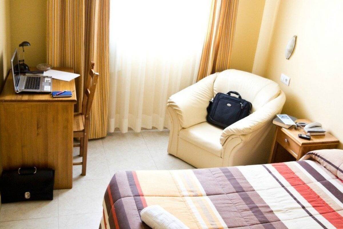Habitación de hotel con cama, escritorio con computadora portátil, silla y mesita de noche con lámpara y bolso.