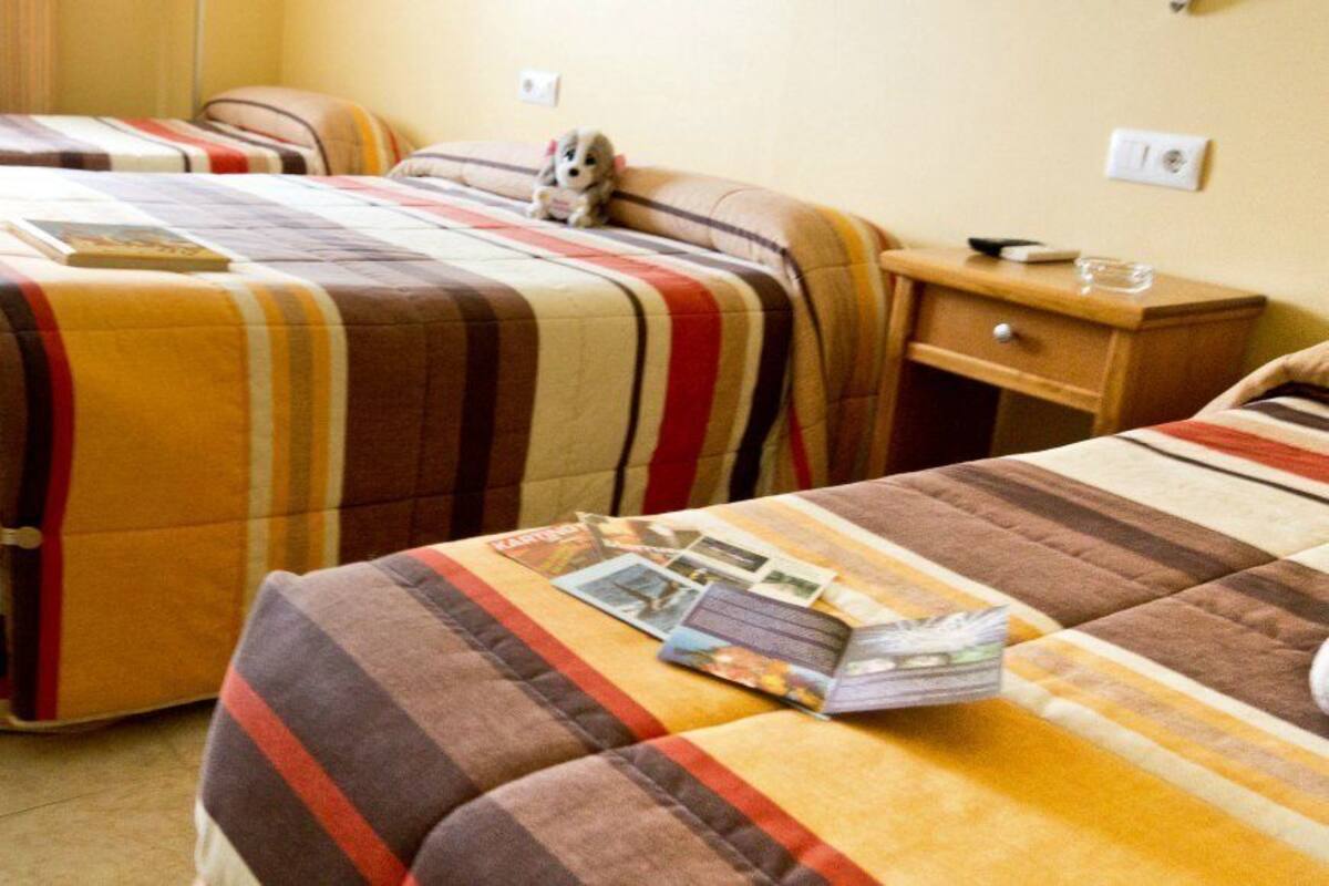 Habitación de hotel con tres camas, cada una con ropa de cama a rayas. Hay una pequeña mesita de noche con una lámpara y un libro.