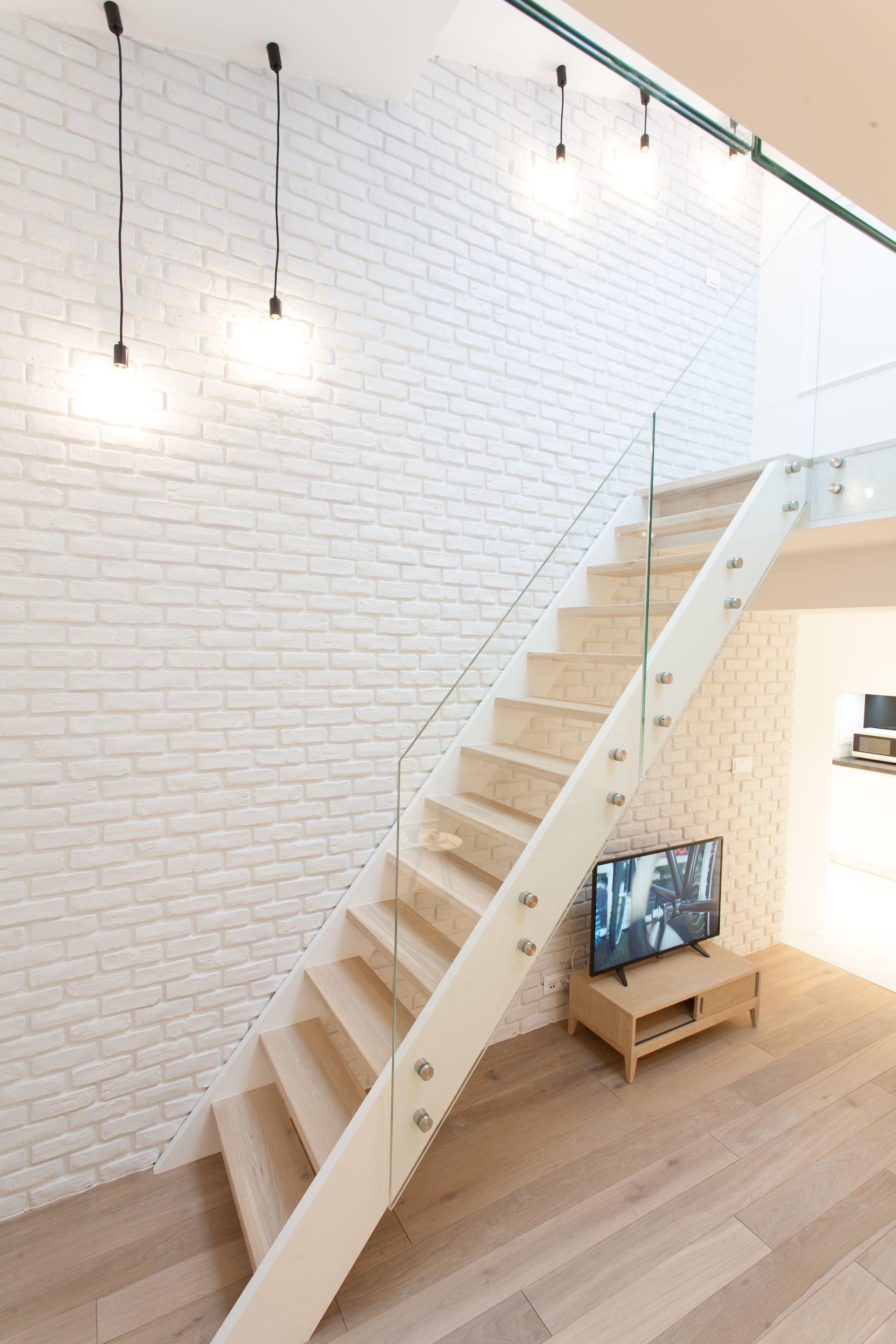 A staircase with a glass railing, a television on a wooden stand, and hanging light fixtures against a white brick wall.