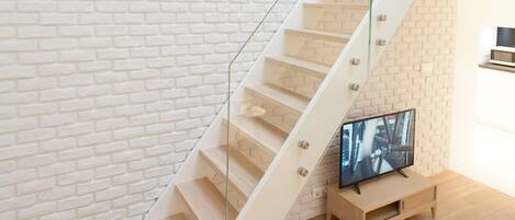 A staircase with a glass railing, a television on a wooden stand, and hanging light fixtures against a white brick wall.