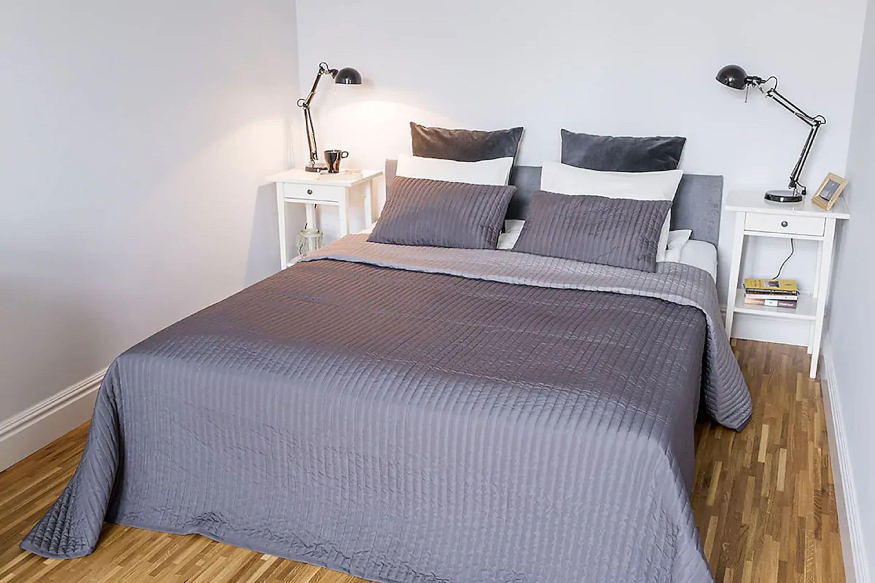 Una cama bien hecha con un edredón gris, dos mesitas de noche con lámparas y un suelo de madera.