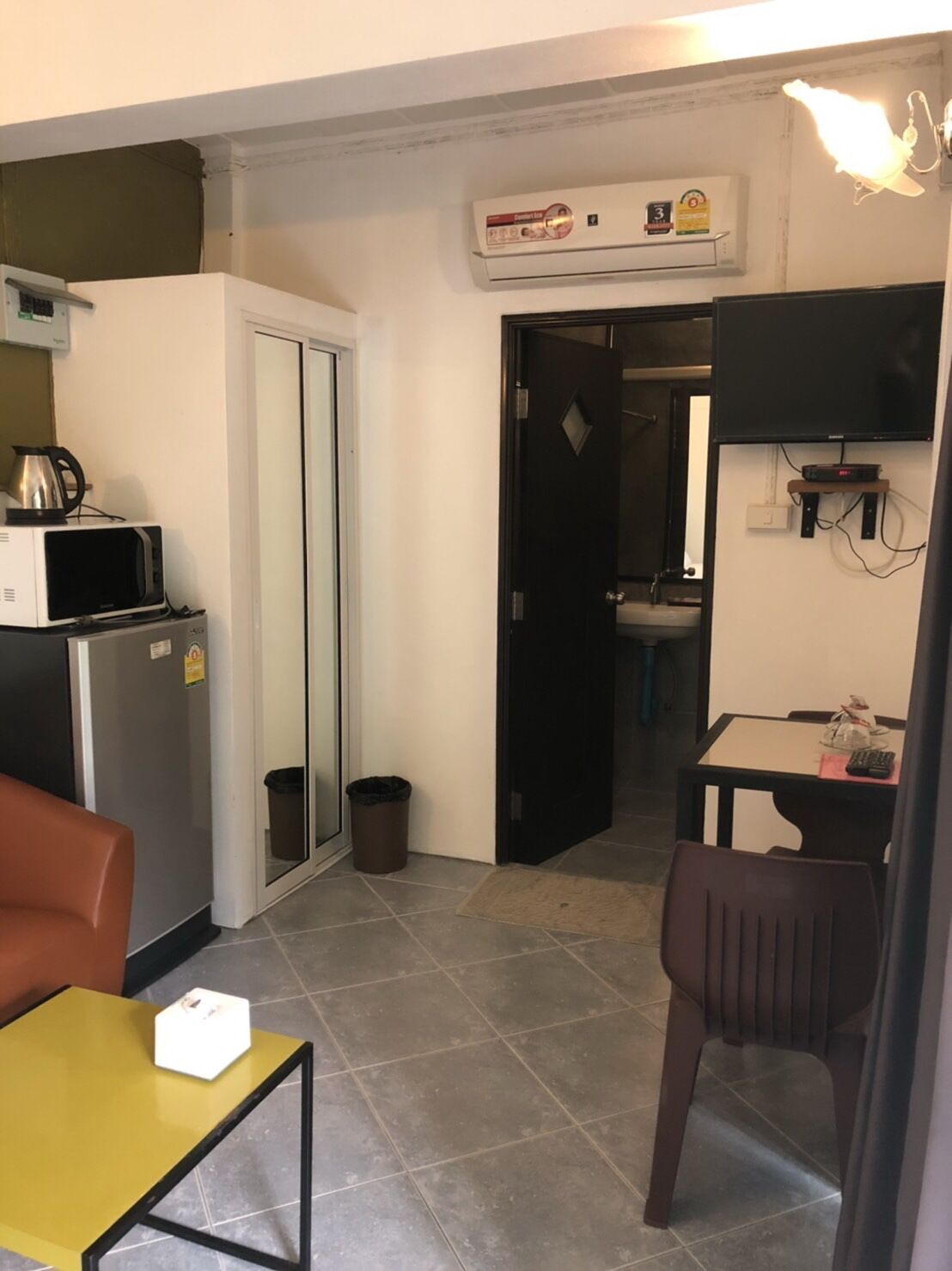 A compact kitchen with a refrigerator, microwave, and coffee maker. There's a small dining table with a chair and a trash bin.