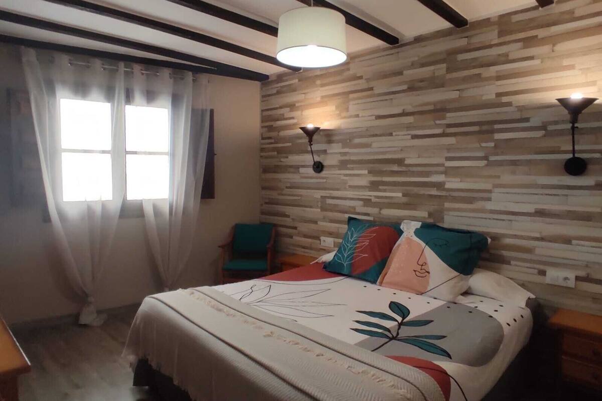 Quarto com cama, janela com cortinas, painel de madeira na parede e duas luminárias fixadas na parede.