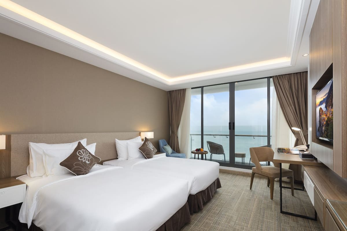 A hotel room with a large bed, a desk with a chair, a TV, and a view of the sea.