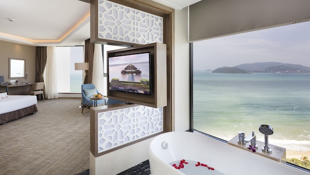 A hotel room with a large window, a TV, a bathtub, and a desk with a chair. - Comodo Nha Trang Hotel (Nha Trang)