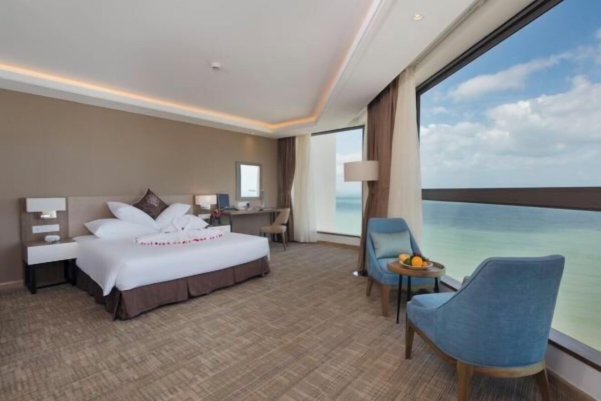 Ein Hotelzimmer mit einem Bett, einem Schreibtisch, einem Stuhl, einem kleinen Tisch mit Früchten und einem großen Fenster mit Meerblick.