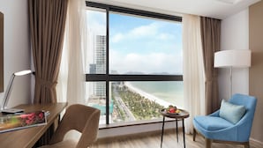 A hotel room with a large window offering a view of a beach and cityscape. There is a desk with a lamp, a chair, and a small table with fruit. - Comodo Nha Trang Hotel (Nha Trang)
