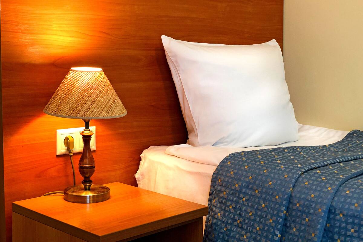 Hotellihuone, jossa on puinen yöpöytä, lamppu, valkoisilla lakanoilla varustettu sänky ja sinivalkoinen kuviollinen peitto.