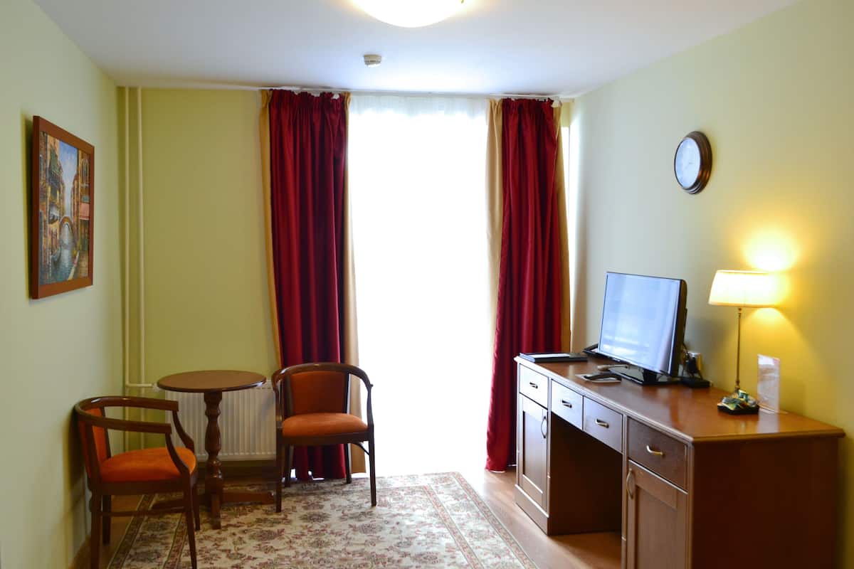Hotellihuoneessa on työpöytä, tuoli ja televisio. Huoneessa on ikkuna, jossa on verhot, patteri ja seinällä oleva kello.