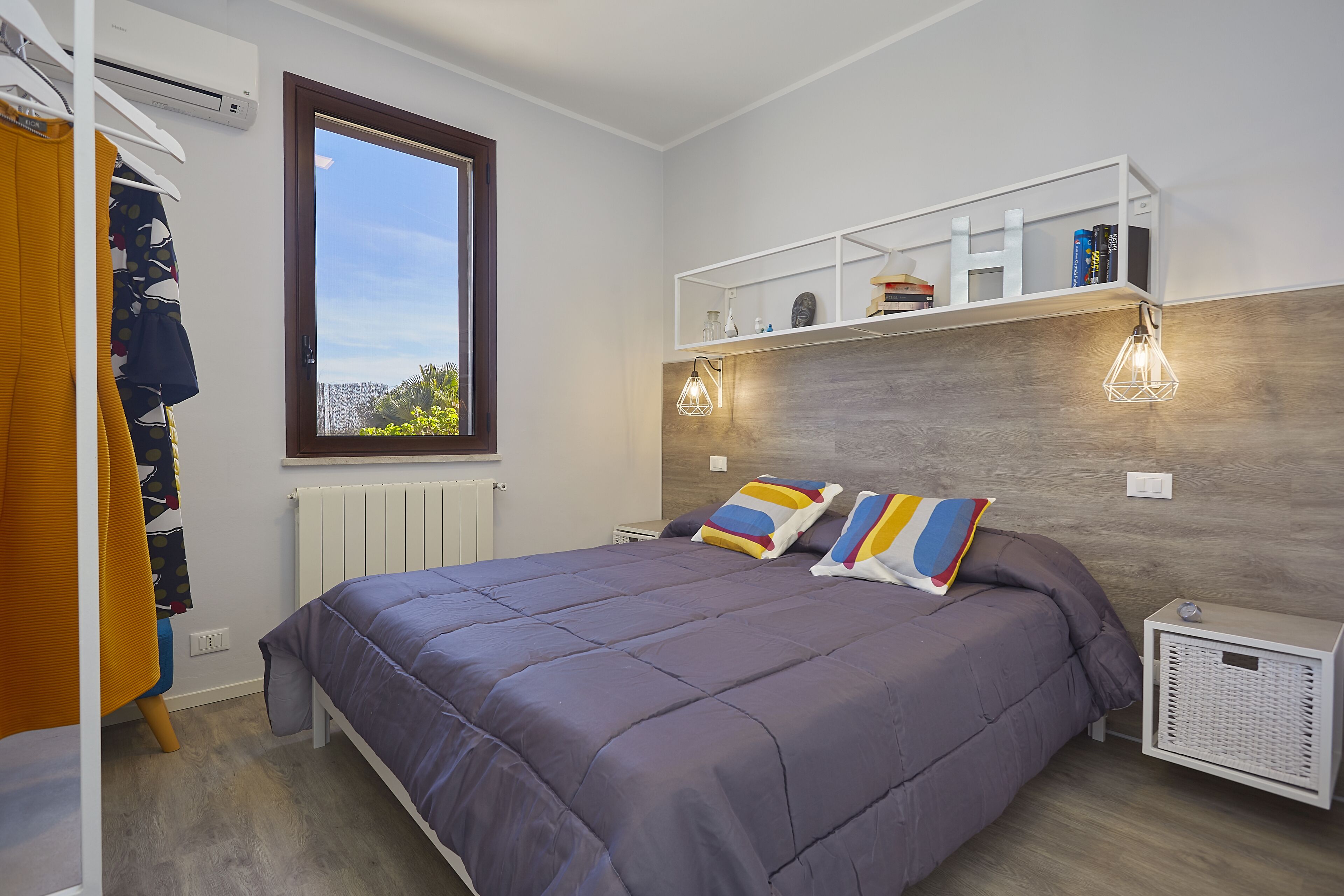 Une chambre avec un grand lit, une armoire, un radiateur, une étagère remplie de livres et une fenêtre donnant sur un beau paysage.