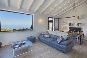 Een moderne woonkamer met een groot raam, een blauwe bank, een eettafel en een keukenhoek.