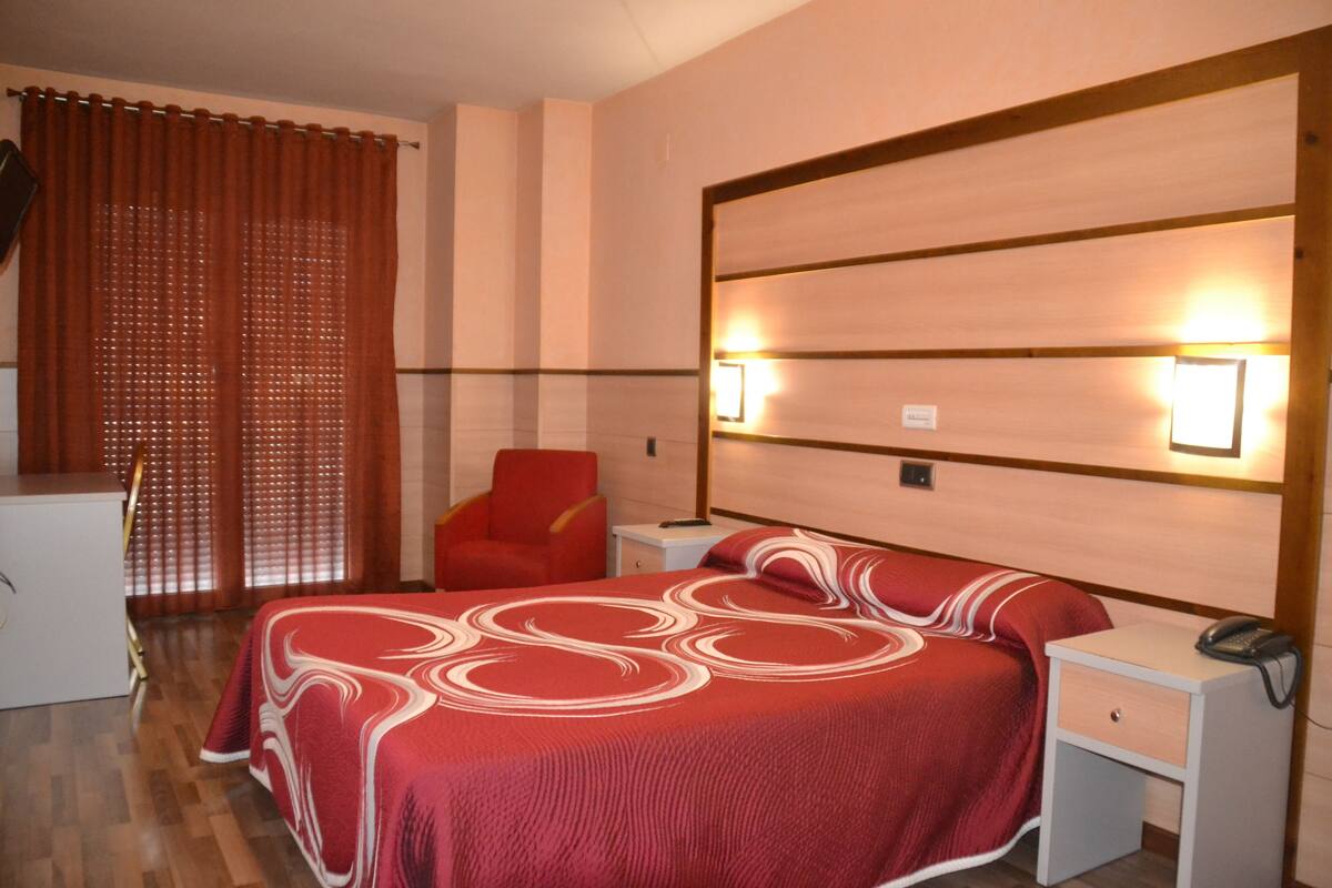 Quarto de hotel com cama, escrivaninha, cadeira e mesa de cabeceira com telefone.
